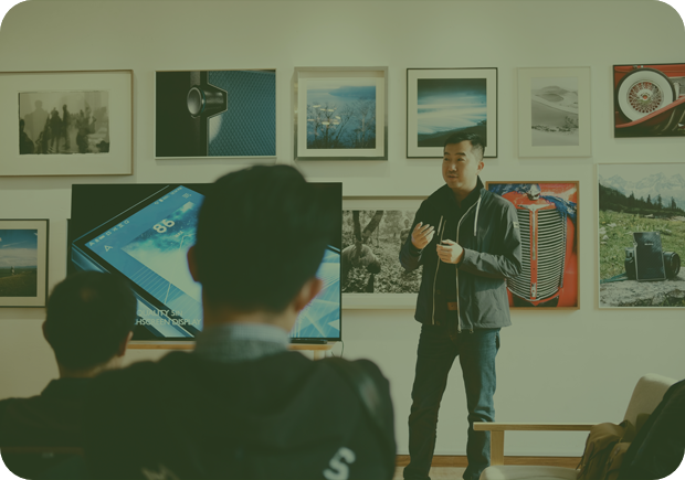 A man, standing in front of a wall of framed photographs, giving a presentation in front of a seated group of people.